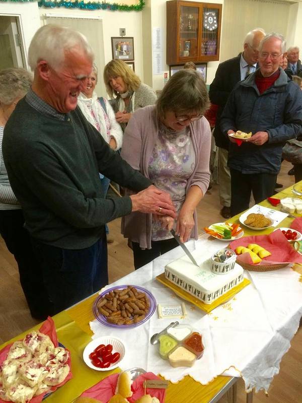 Treve and Gill James cutting a cake, surrounded by well-wishers.