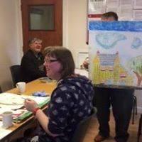 Adrian holding up a different painting, which is obscuring their face. Kelly sat in front working on a painting.